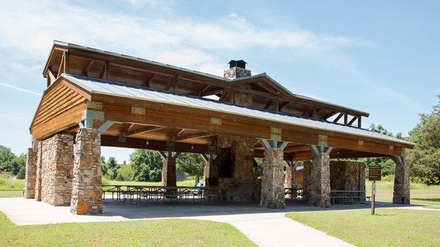 A large open-air picnic pavilion stands in a grassy park under a light blue sky. The structure has a wooden roof with exposed beams and a metal roof, supported by thick stone columns. Beneath the shelter are several picnic tables arranged on concrete.