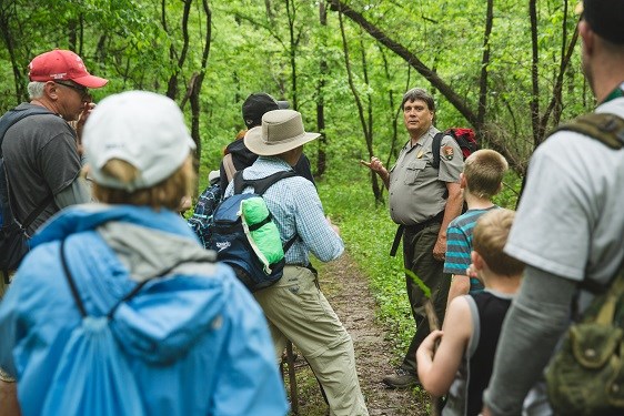 A park ranger leads a group of people down a dirt path through a forest of green trees.