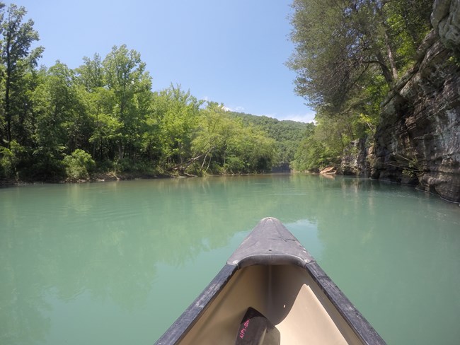 We paddle a canoe across turquoise water with a bluff on our right.
