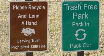 left: brown sign with handshake graphic Lend us a Hand, recycle text; right green sign with recycle symbol graphic, Trash Free Park text