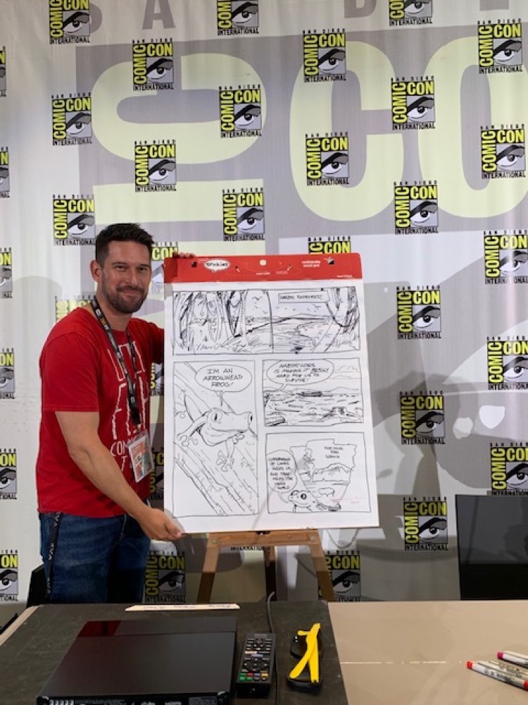 Alonso_Comic-Con Panel 2019 Alonso Nunez from Little Fish Comic Book studio poses next to an easel and large pad of paper with his live-draw, one-page comic on it. The sharpie-drawn comic images depict the Amazon Rainforest and an Arrow Frog.