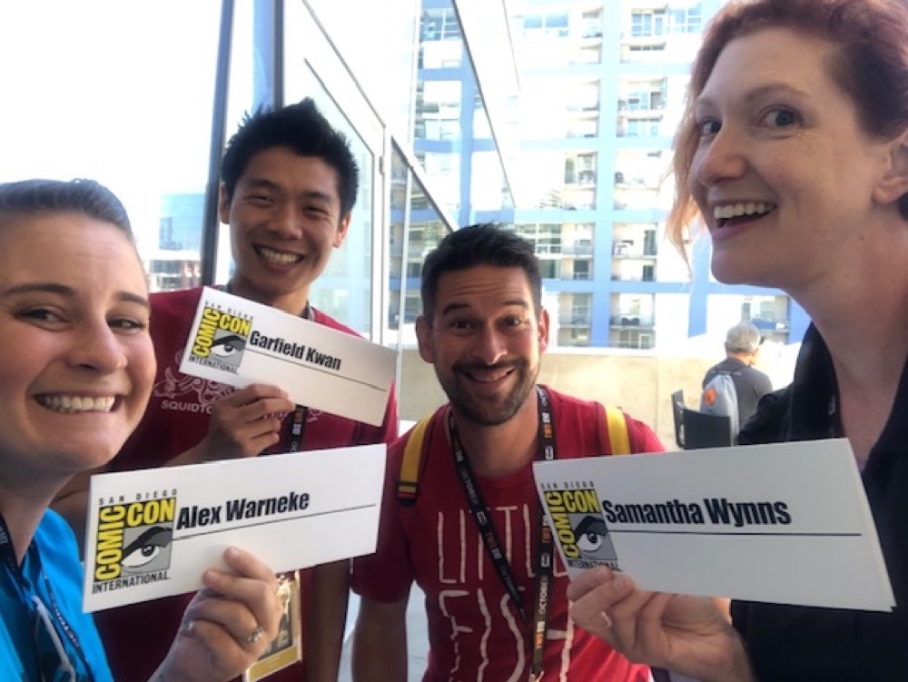 The close-up, grinning faces of Alex Warneke, Garfield Kwan, Alonso Nunez, and Samantha Wynns (left to right) as they hold up their SDCCI nametags after the panel.