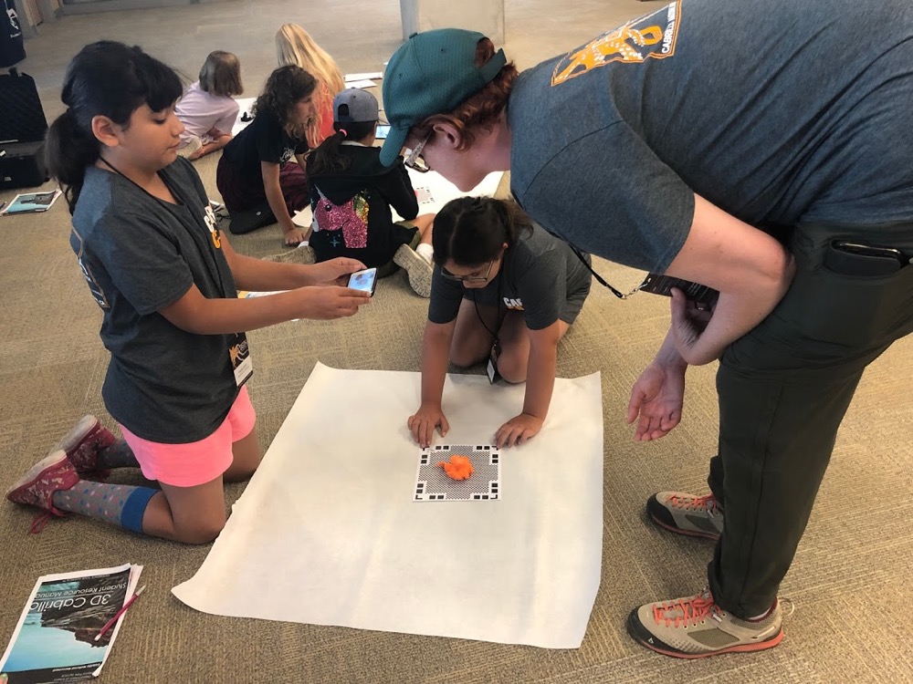 Two EcoLogik campers sit on the carpet to capture images from a stationary object they want to 3D print. An EcoLogik instructor looks on.