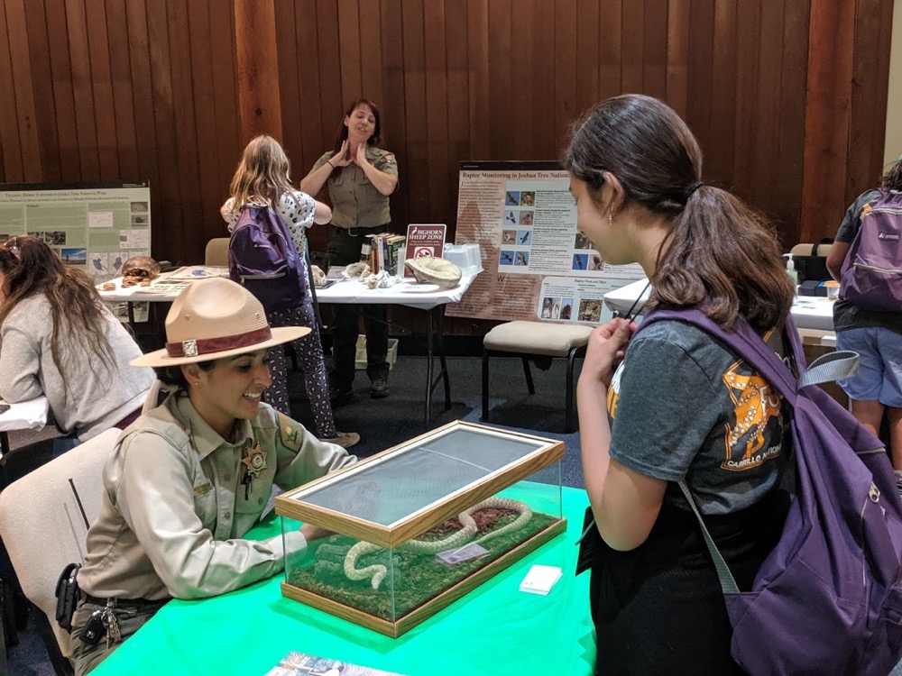 A female County Parks Ranger talks energetically about the snake in the terrarium before her with an EcoLogik camper that stands opposite.