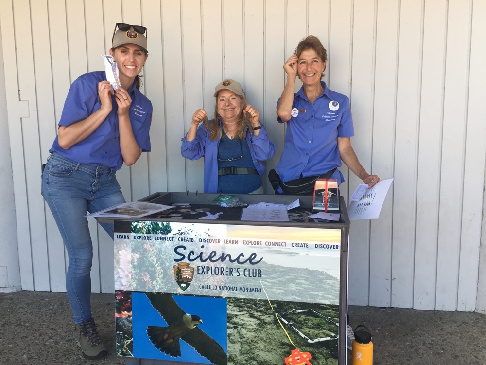 three volunteers stationed at the Science Explorer’s Club table smiling for the camera.