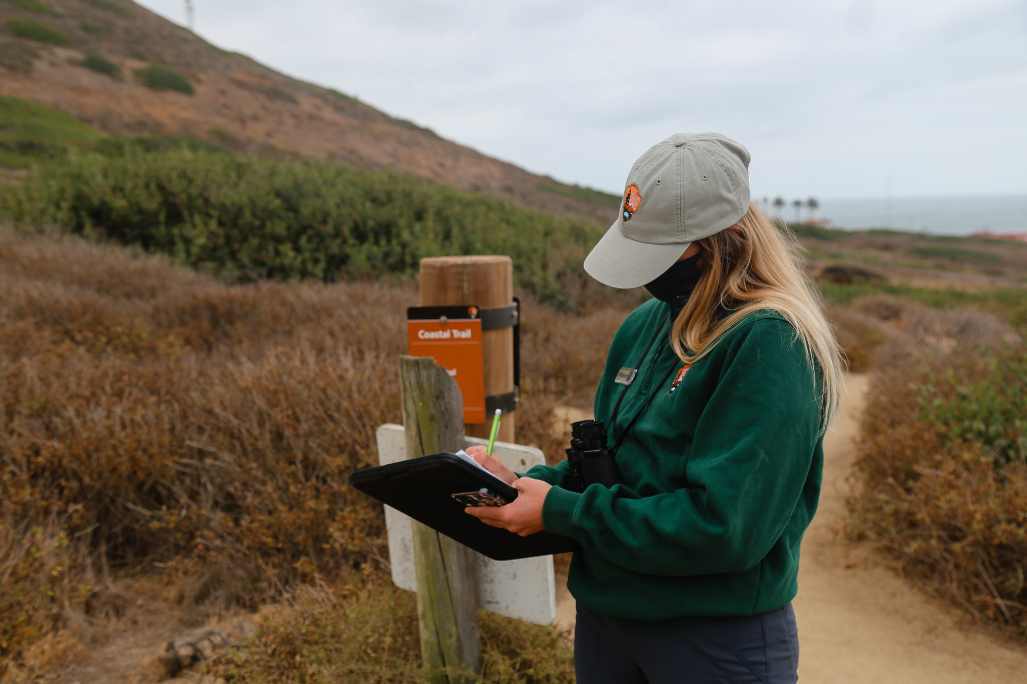 Blonde female wearing a National Park Service sweater and hat writing on a clip board. The woman stands on a dirt path that runs into the background. 