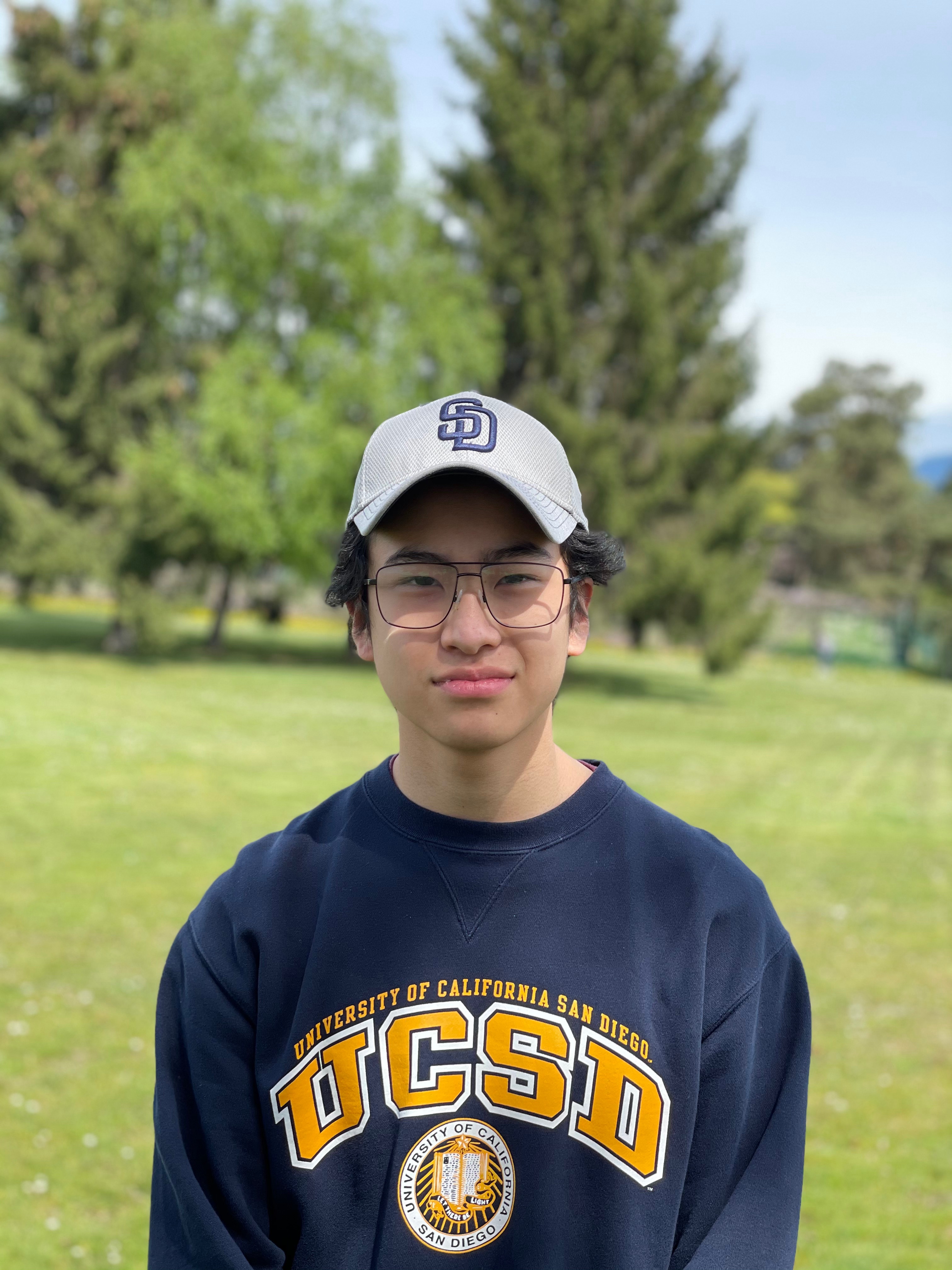 A man wearing glasses, a blue sweatshirt with gold letters stating “UCSD”, and a gray ballcap with “SD” embroidered on it stands on a green field. 