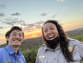Keanu on the left and Ranger Sandy on the right, smiling with the hills of Cabrillo National Monument. A yellow and orange sunset can be seen settling over the hills.