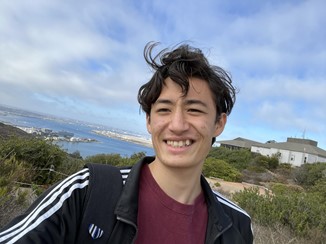 Keanu smiling with wavy hair swept by the ocean breeze with part of the Visitor Center and San Diego Bay in the background. 