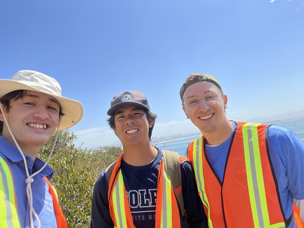 : Interns Keanu, Taro, and Brent stand alongside each other in orange and yellow safety vests with the view of San Diego’s bay in the background. Names go left to right in standing position.