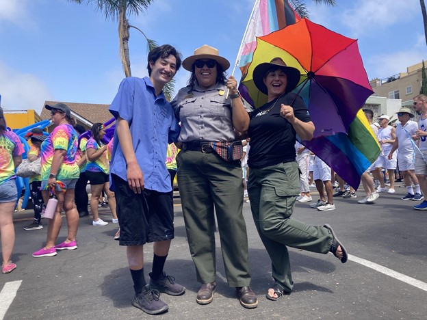 Intern Keanu, Ranger Sandy, and Greenhouse Manager Patricia holding arms. smiling with groups of people in tie-dye and white shirts in the background. Patricia holds a rainbow parasol with one leg up, Sandy in uniform, and Keanu in volunteer uniform.