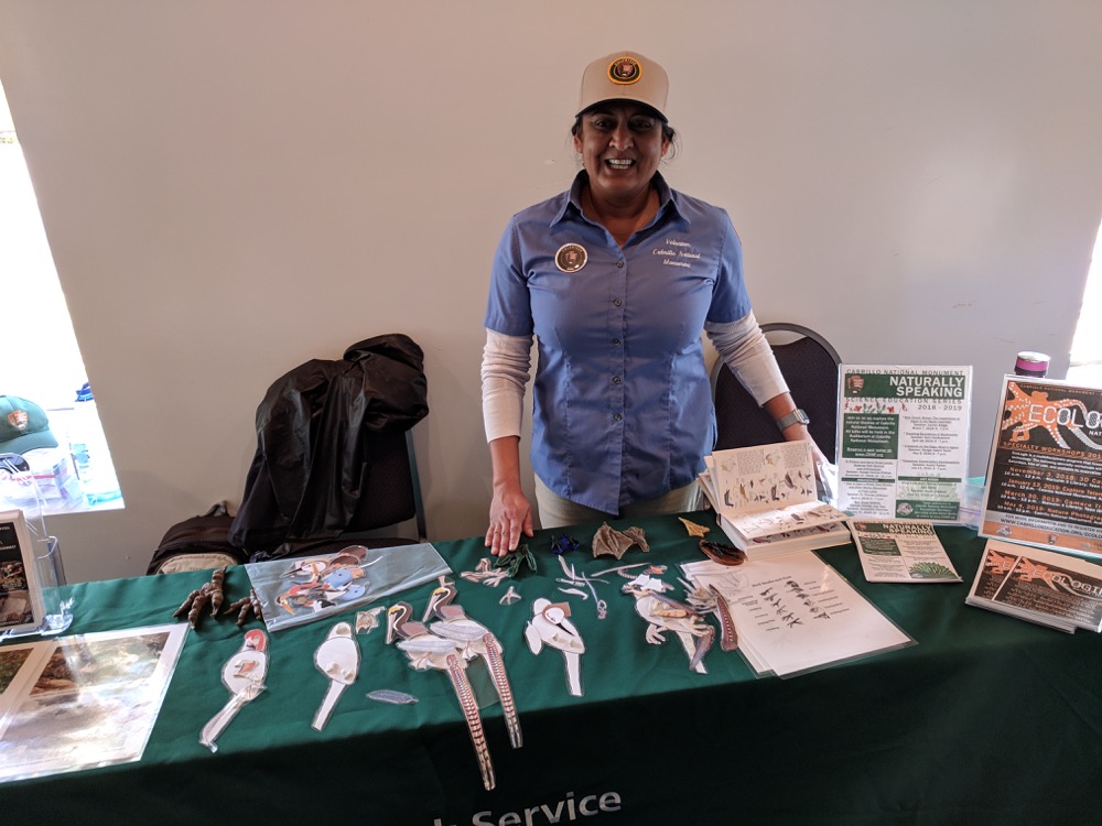 Volunteer Alida stands smiling behind a table that displays the Build-a-Bird activity and promotional materials. 