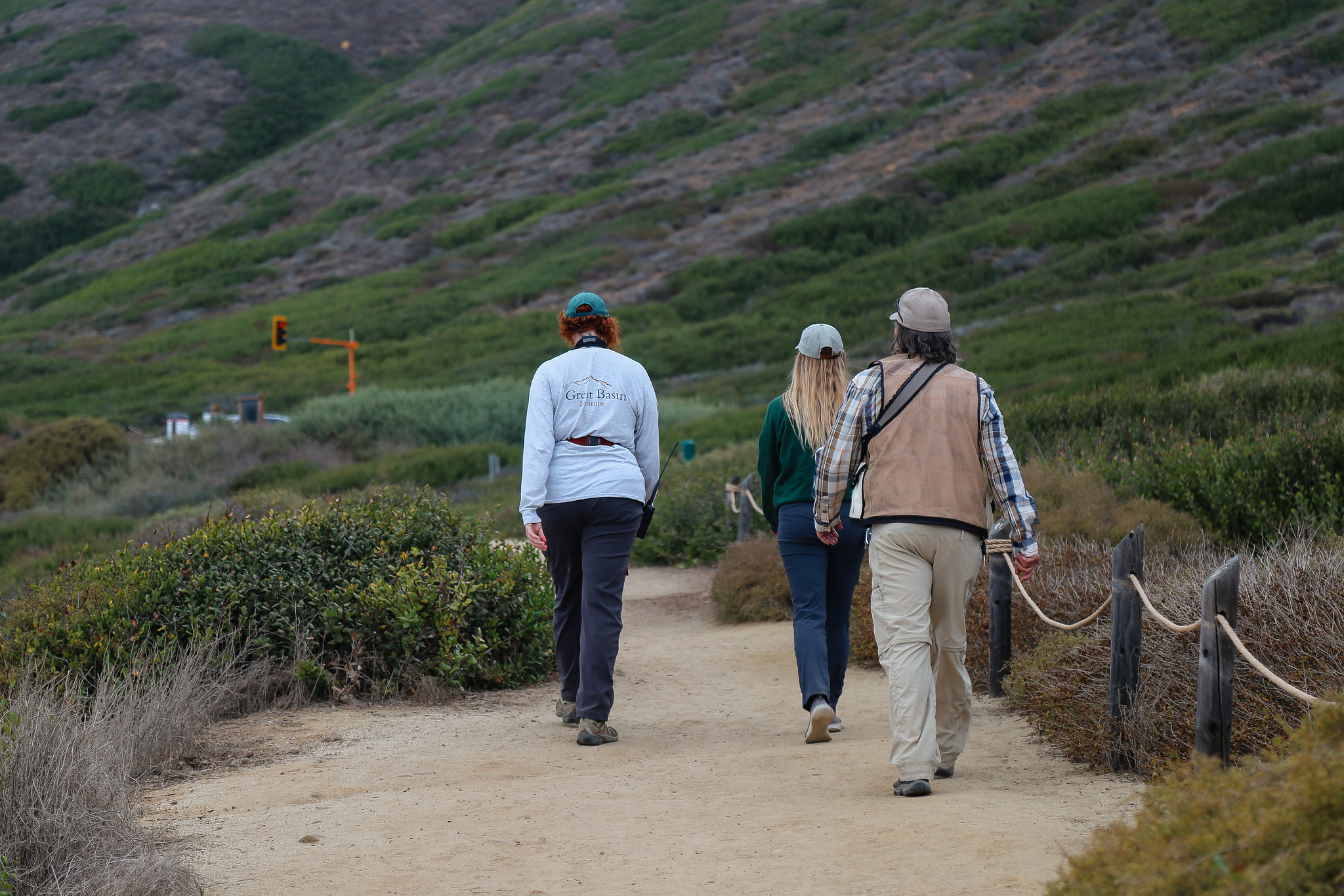 Two women and one man walking down a dirt path with coastal sage scrub plants lining the trail on both sides. 
