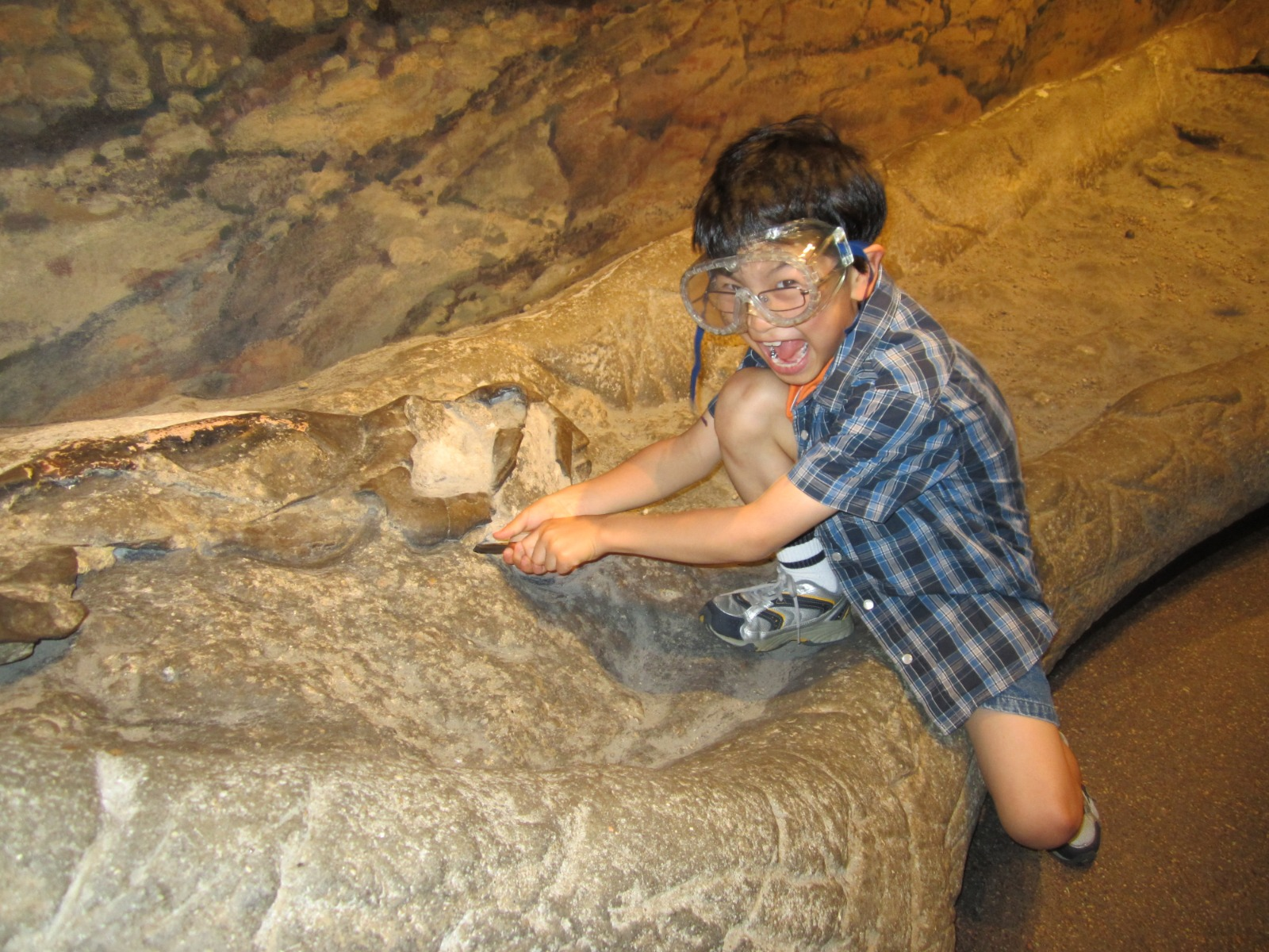 a young Kazune wearing a blue checkered shirt and safety goggles crouches excitedly on a large fossil, pretending to dig it up. 