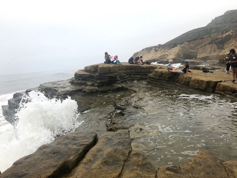 a wave crashes onto the cliffs near some students at Cabrillo National Monument.