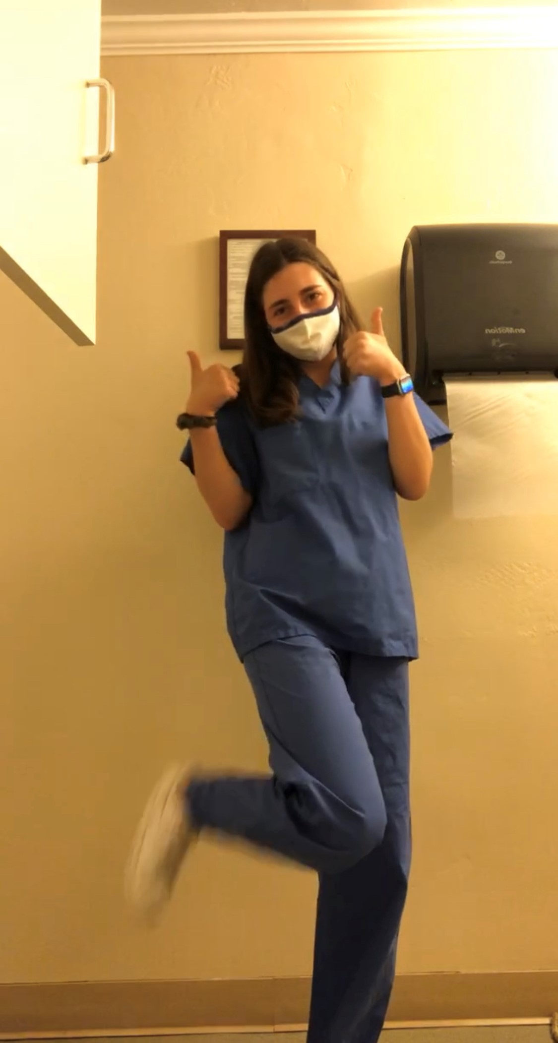 A person standing in a medical office with long brown hair wears blue medical scrubs, a white facemask, and white sneakers. They give the camera two thumbs-up.