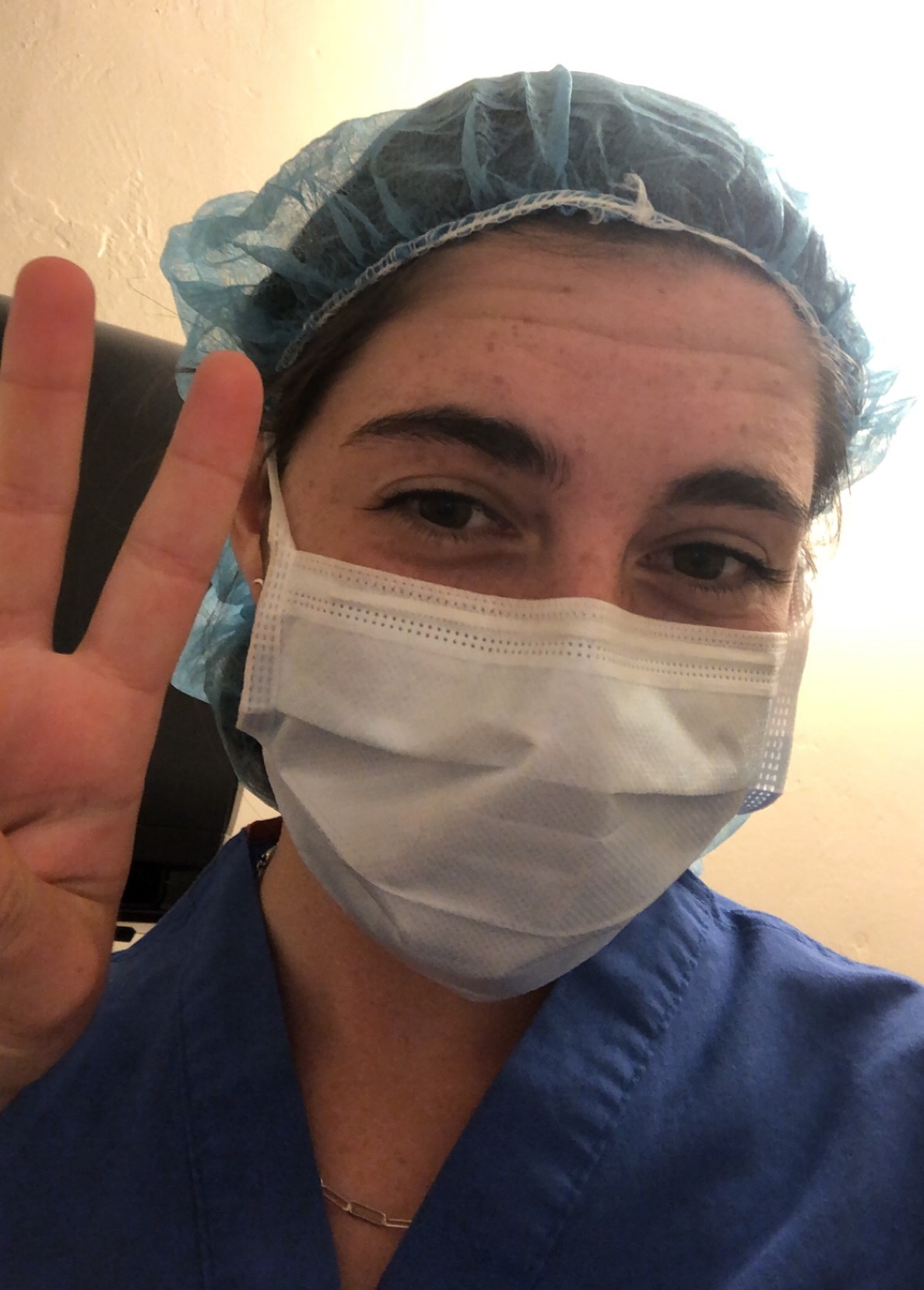 A close-up of a person with brown eyes and freckles who’s wearing blue scrubs, a white medical facemask, and a blue surgical scrub cap. They hold up two fingers in a “peace” sign.
