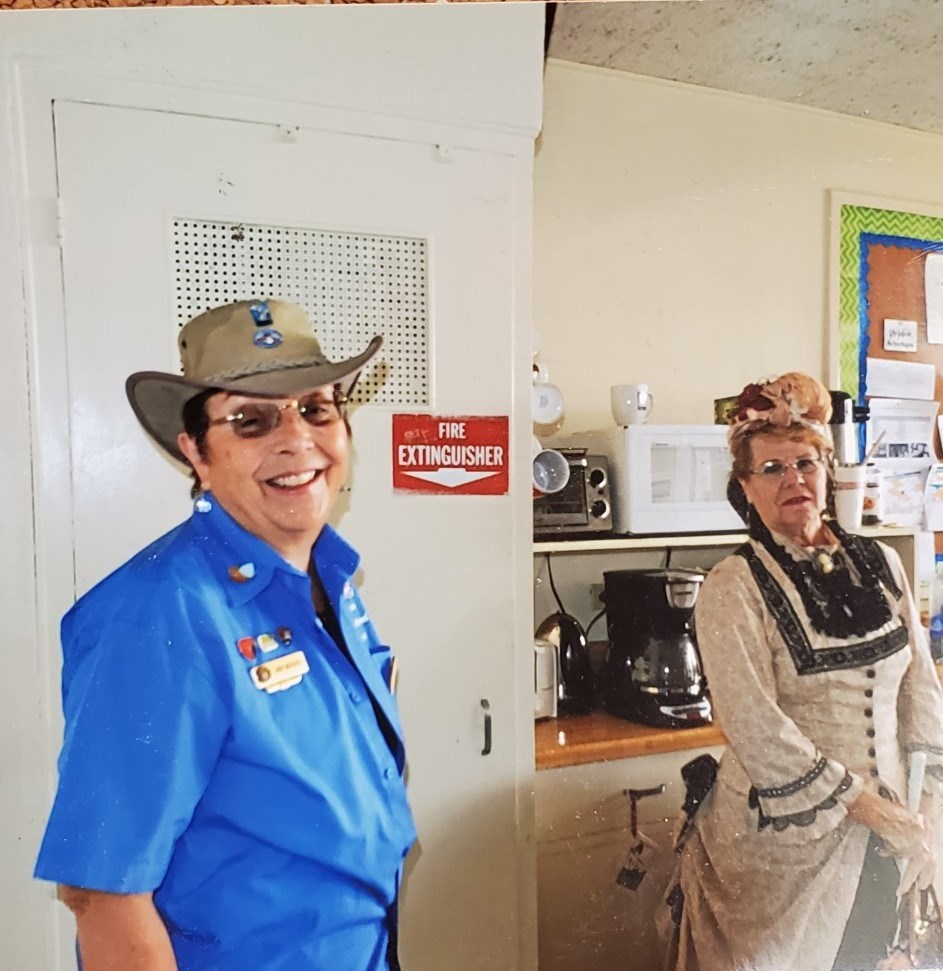 Two women, one in a blue volunteer shirt and tan hat (left) and the other in a 1800s-style dress (right) smile and stand in an office kitchen.