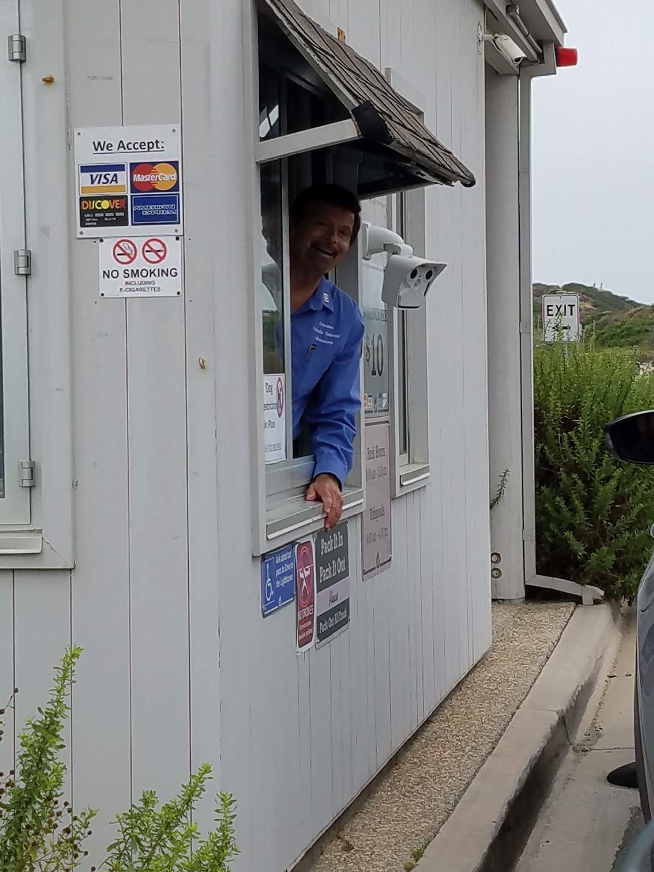 A volunteer in a blue volunteer shirt peeks his upper torso out of a white entrance station’s window, surrounded by an assortment of signs.