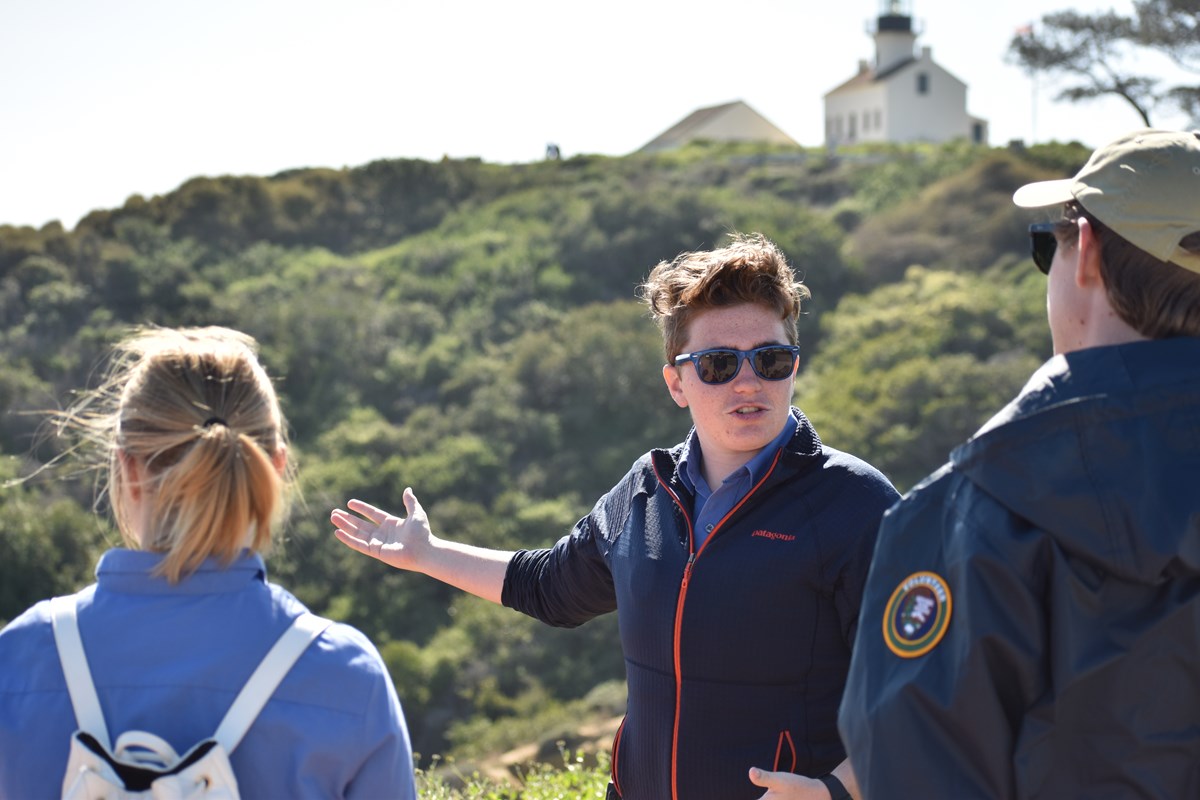 Two volunteers, one in a blue volunteer shirt and white backpack (left) and the other in a navy-blue volunteer windbreaker stand facing another volunteer tour guide (middle) who gestures to the right. Behind them is a white lighthouse structure.