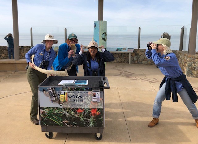 Three people in blue uniforms, stand holding whale bones and baleen behind a roll-away cart, while a third person stands facing them off to the side looking through binoculars. Behind them is an ocean lookout with a stone wall and glass screens.