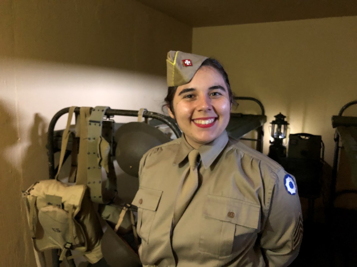 A woman in a WWII military uniform stands and smiles inside a military housing room, which includes bunk beds, backpacks, helmets, and a lantern.