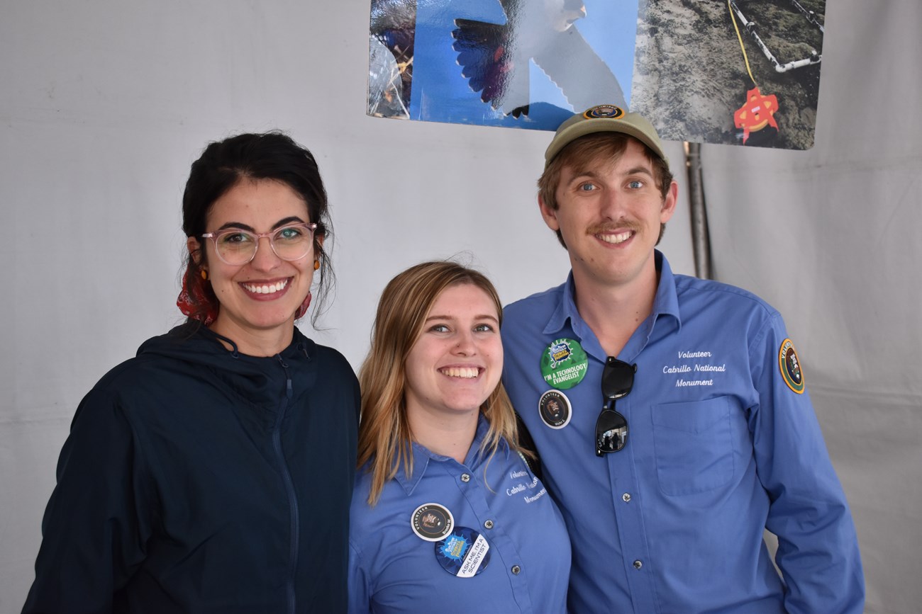 Three people—one woman in a navy-blue windbreaker (left), another woman in a blue volunteer shirt (middle), and a man in another blue volunteer shirt (right)—stand next to a white tent and smile. Behind them is a hanging poster of various nature shots.