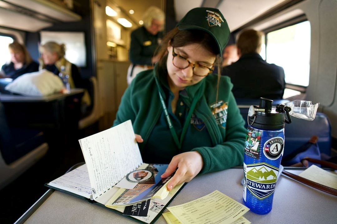 A woman in glasses and a green, volunteer outfit logos sits inside of a train and looks inside a journal, filled with notes and loose brochures, that sits on a table beside a bright blue water bottle with an assortment of stickers and other yellow papers.
