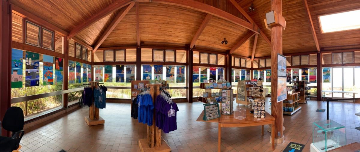 Colorful student art on squares canvas covers the windows in a large building with wooden ceilings. Store shelves are covered with gifts and books.