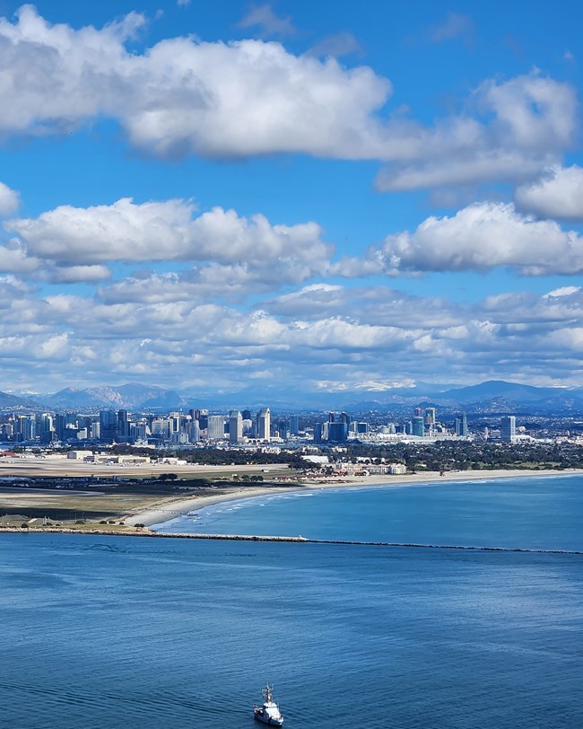 A closer view of the beaches, skyscrapers, and mountains in the San Diego skyline as seen from Cabrillo National Monument.  Up front, a small boat goes by the bay.