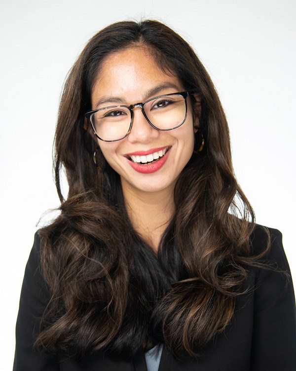 Smiling woman with long, wavy brown hair wearing black blazer and glasses.