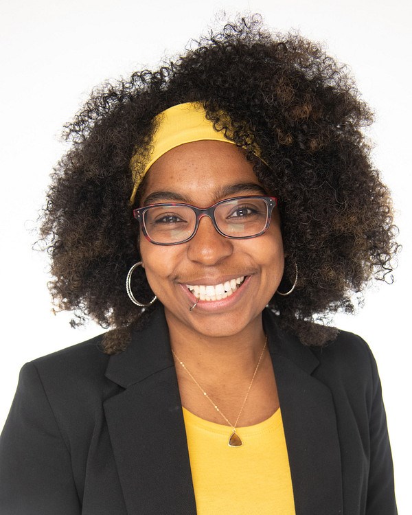 Smiling woman with brown afro wearing glasses, black blazer, yellow shirt.