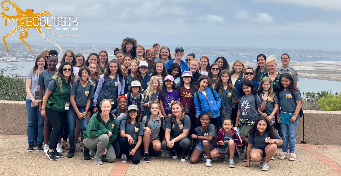 Group of women and girls pose for picture with San Diego Bay and the city in the background. In the corner is the "EcoLogik Nature x Tech" logo.