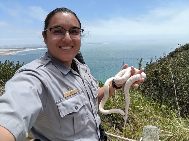 A woman with dark hair and glasses holds a snake in her hand. The ocean is in the background.