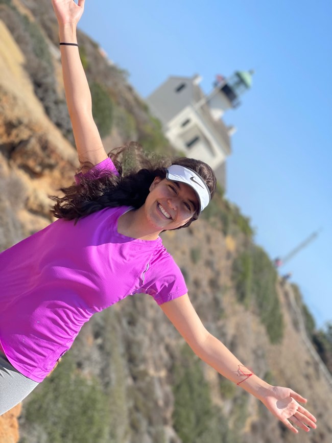 A woman in pink shirt and white visor has arms extended. Lighthouse is in background.