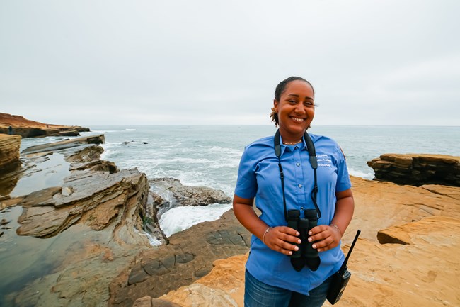 A woman in a blue shirt with binoculars around her neck stands on cliff overlooking the ocean.