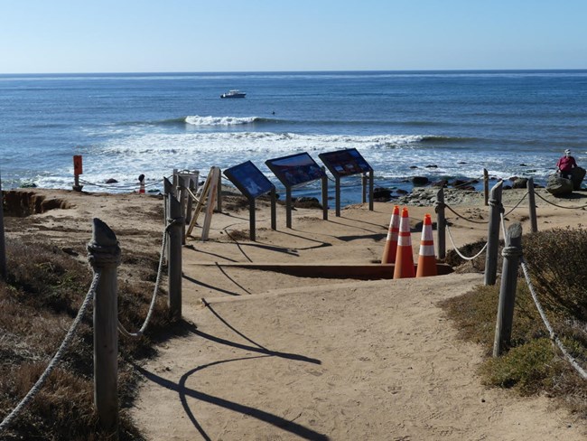 A dirt path with wooden posts and rope along the sides leads to the ocean and the tidepools. Three waysides are on the left side along the path.