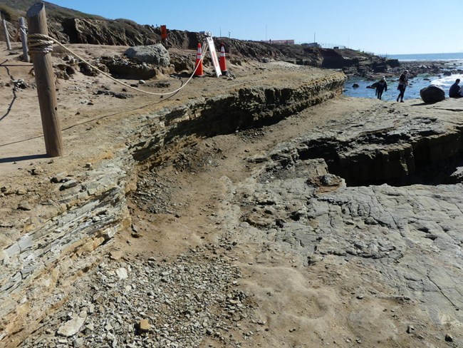 A narrow rocky and dirt path along rocky sandstone cliffs lead the way to the tidepools. Post and rope are along the cliff edge.