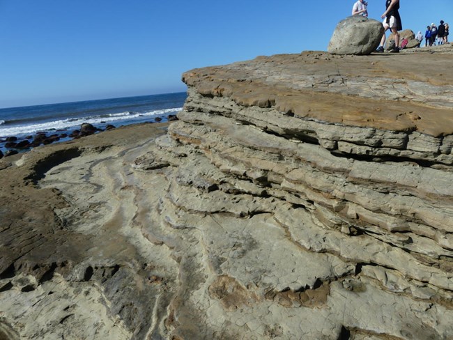 Cliff ledges formed by soft sandstone layers. Large boulders are at the top of the cliff.