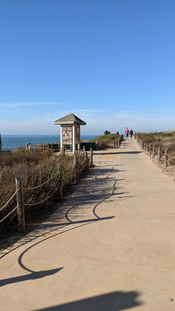 A dirt path with low lying shrubs on both sides. Wooden post and rope line the path. A kiosk is ahead on the left.