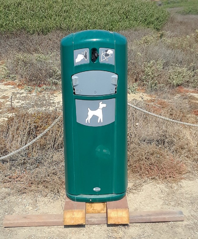 Green enclosed metal can with logo and hole for bags at top and logo and hole for trash at bottom.