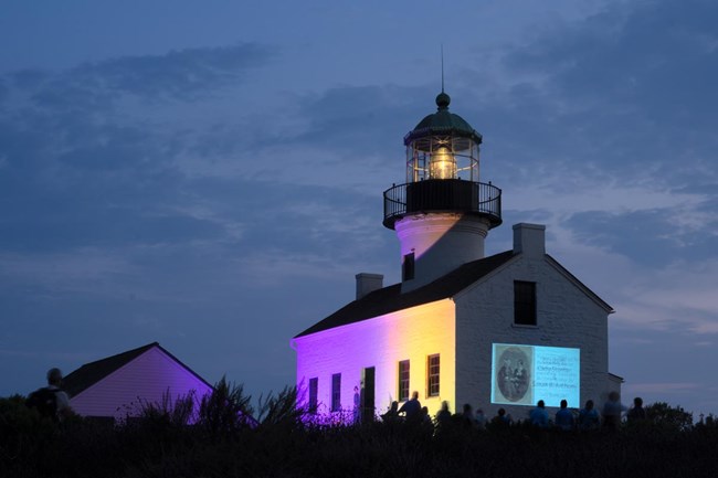 White two-story lighthouse with lit lantern in top tower and front lit in purple and yellow lights. A historic image of two women is projected on a side wall and people are gathered around to look at it. The sky is blue and dark.