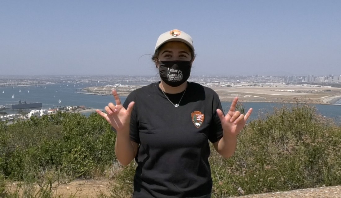 AS LTours Woman in hat and shirt with National Park Service arrowhead logo holds up both hands with thumb, pointer finger, and pinky finger extended. Behind her a waterway with boats and city skyline are visible.
