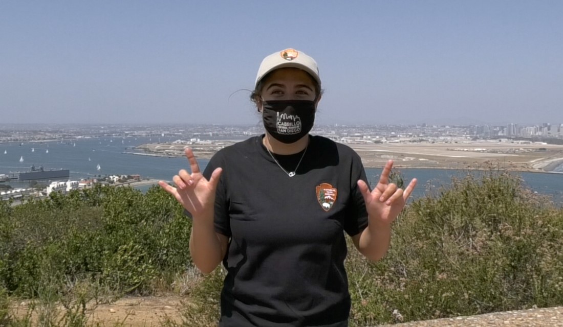 Woman in hat and shirt with National Park Service arrowhead logo holds up both hands with thumb, pointer finger, and pinky finger extended. Behind her a waterway with boats and city skyline are visible.