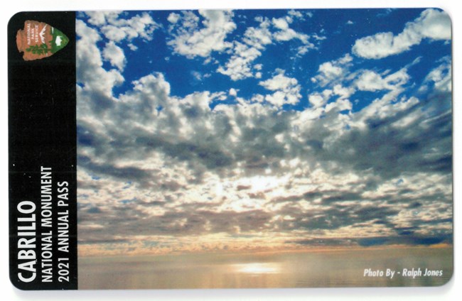 2021 Cabrillo Annual Pass. Depicts patchy clouds hanging over silver-gold water, beneath a blue sky.