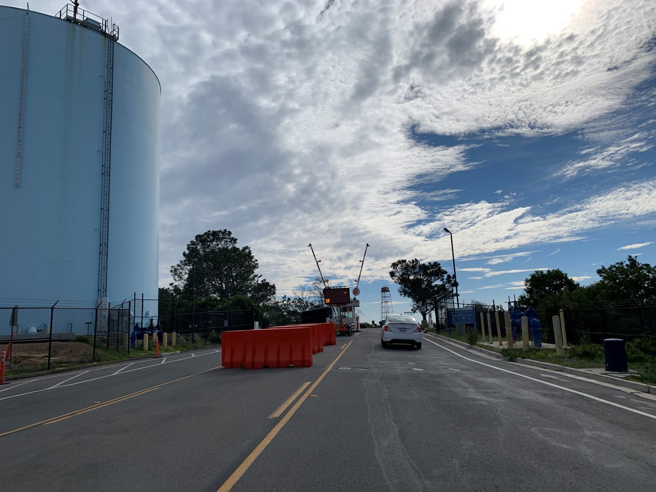 A car driving down a road and stopping at a small white building with gates across the road. A large blue tank is on the left side of the road.