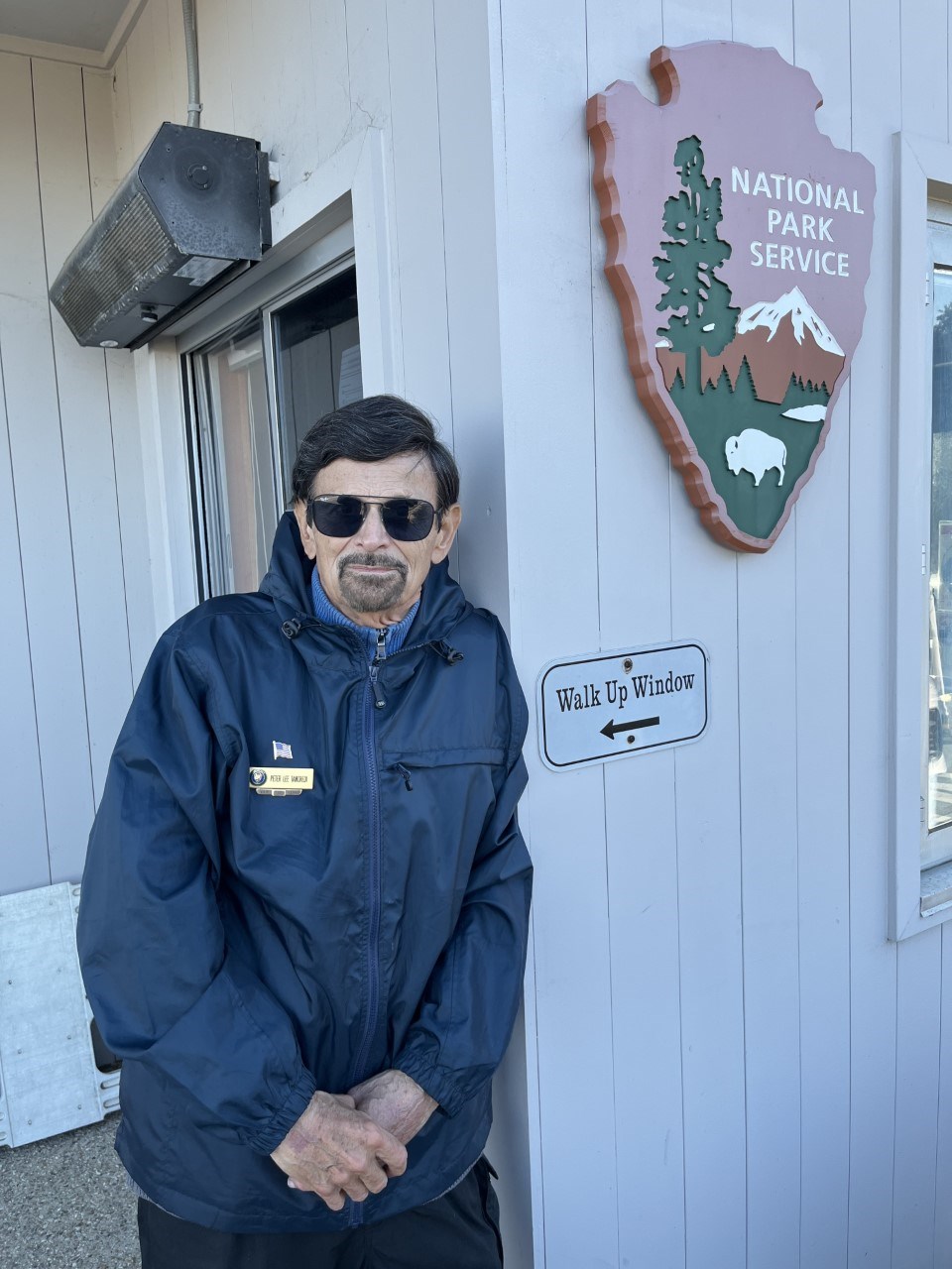 A park volunteer wearing a blue jacket and sunglasses stands next to a park service arrowhead sign and a small sign that reads “Walk up Window”