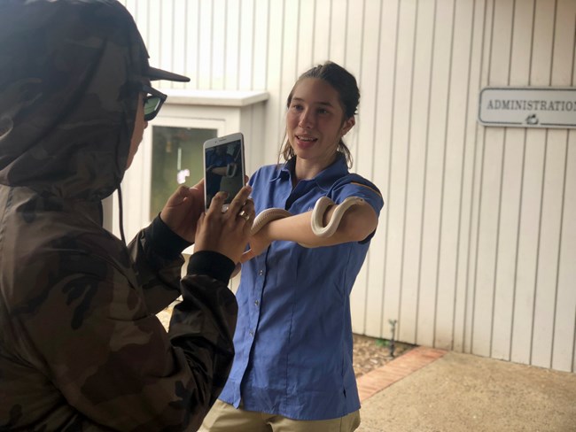 A smiling woman in a blue shirt with a white snake coiled around her arm stands talking to a person who takes a picture of the woman with their phone.