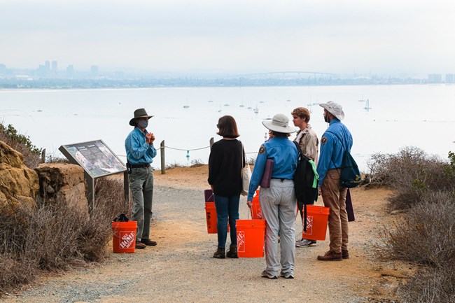 Weed Warrior volunteers gather around park staff to learn about invasive plants on Bayside Trail.  They hold orange buckets. In the background is the San Diego Bay and city skyline.