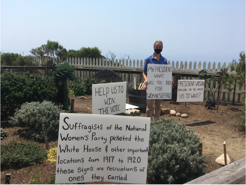 White signs with black text and metal stakes inside a garden with wooden fence. A young woman holds up one of the signs.